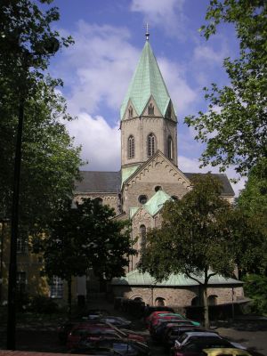 Abgebildet ist ein Foto der Kirche Basilika Sankt Ludgerus. Im Vordergrund stehen einige Autos auf einem Parkplatz und an den beiden Bildrändern sind einige größere Bäume zu erkennen. In der Bildmitte ist die aus Bruchsteinen gemauerte Kirche mit einem achteckigen Turm zu erkennen.